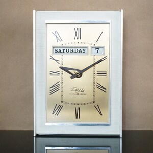 Puede incluir: Un reloj vintage General Electric Date Mate blanco con una esfera dorada y marcadores de hora romanos negros. El reloj muestra la fecha "7" y el día "SÁBADO".