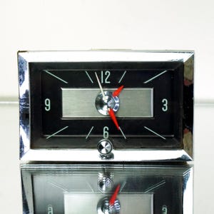May include: A rectangular clock with a black face, silver accents, and a chrome frame. The clock has white numerals at the 3, 6, 9, and 12 positions, with red hands. The clock is reflected in the surface below.