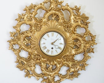 May include: A gold-toned wall clock with an ornate, decorative frame. The clock face is white with black Roman numeral hour markers and black hands.