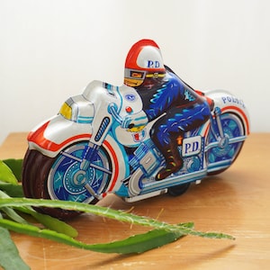 Könnte beinhalten: Ein weißes und blaues Aufziehspielzeug-Motorrad mit einer Polizeifigur, die darauf sitzt. Das Motorrad hat einen roten Streifen und die Aufschriften "Police" und "P.D." darauf. Der Polizist trägt eine blaue Uniform und einen weißen Helm mit "P.D." darauf.