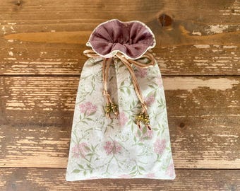 Puede incluir: Una bolsa de tela con cordón ajustable que presenta un exterior claro con un delicado estampado floral de flores rosas y hojas verdes. El forro interior es de color malva sólido y la bolsa se cierra con cordones finos terminados en pequeñas cuentas metálicas.