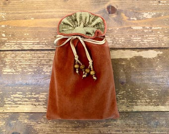 Puede incluir: Una bolsa de terciopelo color óxido con una parte superior con volantes dorada y verde oliva. El cordón es dorado con cuentas de madera. La bolsa está sobre una superficie de madera.