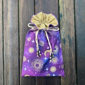 Könnte beinhalten: Lilafarbener Kordelzugbeutel mit einem goldenen und weißen Himmelsdruck. Die Tasche hat ein goldenes Stofffutter und einen perlenbesetzten Kordelzug.