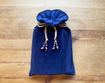 May include: A navy blue velvet drawstring bag with a gold-coloured cord and beaded tassels. The bag is closed, with the top gathered and tied. The background is a light brown wooden surface.