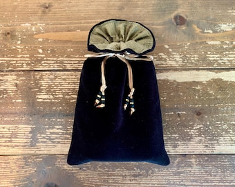 Puede incluir: Una bolsa de terciopelo negro con cordón y forro interior dorado metalizado. La bolsa se cierra en la parte superior con un cordón fino de color claro que termina en dos borlas decorativas, adornadas con pequeñas cuentas oscuras y detalles dorados.