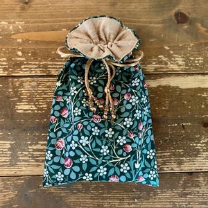 Puede incluir: Una bolsa con cordón con un estampado floral en verde oscuro, blanco y rosa. La bolsa tiene una parte superior beige fruncida y un cordón dorado con cuentas decorativas. La bolsa está sobre una superficie de madera.