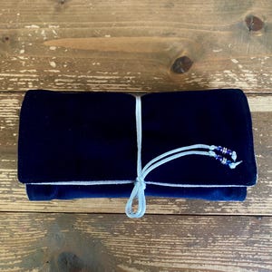 May include: A navy blue velvet roll-up case tied with a light blue cord. The case is rectangular and appears to be closed. The cord is adorned with small, dark blue beads. The case is resting on a wooden surface.