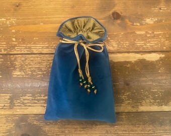 Puede incluir: Una bolsa de terciopelo azul profundo con cierre de cordón, que presenta un forro interior de color oro metálico y un cordón dorado. Los extremos del cordón están decorados con pequeñas cuentas cilíndricas de color verde oscuro.