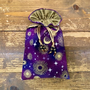 Puede incluir: Una bolsa morada con cordón y un diseño celestial con rayos de sol, lunas y estrellas doradas. La bolsa tiene una parte superior dorada con volantes y cordones dorados con borlas de cuentas negras. La bolsa está sobre un fondo de madera.