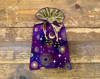 May include: A purple drawstring bag with a celestial design featuring gold sunbursts, moons, and stars. The bag has a gold-coloured, ruffled top and gold drawstring cords with black beaded tassels. The bag is set against a wooden background.