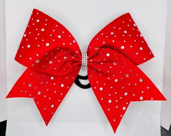 May include: A bright red hair bow made of grosgrain ribbon, featuring a center knot embellished with clear rhinestones. The entire surface of the bow is decorated with scattered clear rhinestones of varying sizes, and a black elastic hair tie is attached.