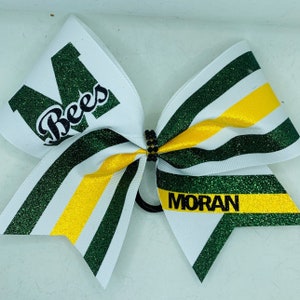 May include: A white cheer bow with green and yellow stripes. The bow has a black letter "M" with the word "Bees" in black lettering. The other side of the bow has the word "MORAN" in black lettering.