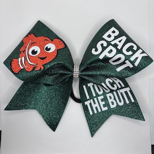 Pode incluir: Laço de animadora verde com glitter, com a inscrição branca "Back Spot I Touch The Butt" e um peixe laranja e branco de desenho animado do outro lado.