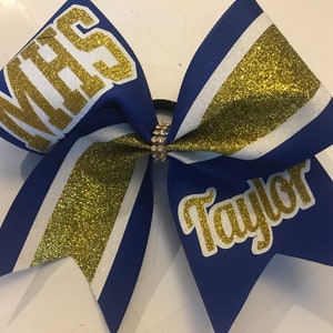 May include: A blue and gold cheerleading bow with the text "MHS" and "Taylor". The bow features gold glitter stripes and a rhinestone center. This accessory is perfect for cheerleaders.