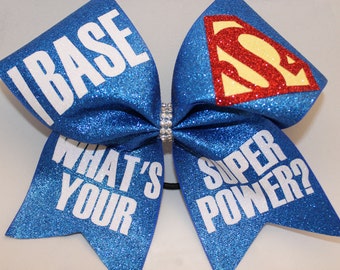 May include: A blue glitter cheerleading bow with white text that reads "I BASE WHAT'S YOUR SUPER POWER?" The bow features a Superman logo in red and yellow. A rhinestone band is in the center.