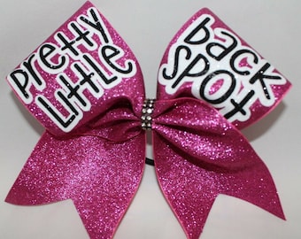 May include: A pink glitter cheer bow with white and black lettering. The left side of the bow says "pretty little" and the right side says "back spot".