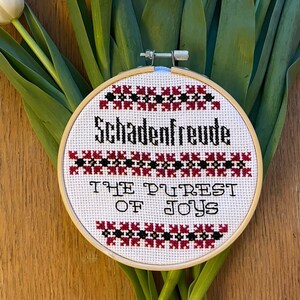 May include: A white embroidery hoop with a red and black border. The hoop features the text "Schadenfreude" and "The Purest of Joys" in black.