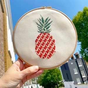 Puede incluir: Bordado de punto de cruz de una piña en un bastidor de madera. La piña presenta un patrón de diamantes rojos y dorados con hojas verdes. El bordado está sobre una tela color crema.