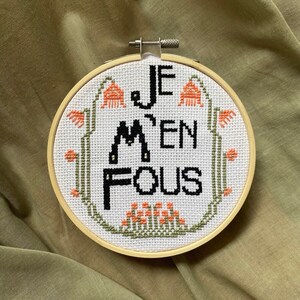 Puede incluir: Un aro de bordado blanco con un diseño de punto de cruz en blanco y negro que dice "Je m'en fous". El diseño está rodeado de pequeñas flores naranjas.