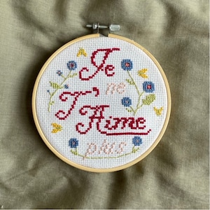 May include: A white embroidery hoop with a red embroidered phrase "Je T'aime" surrounded by blue and yellow flowers.