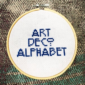 May include: A white embroidery hoop with a blue cross-stitch design that reads "ART DECO ALPHABET".