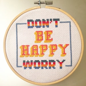 May include: A white embroidery hoop with a cross-stitch design that reads "Don't be happy, worry". The text is in a retro font and is stitched in yellow, orange, red, blue, and black thread.