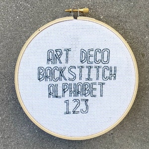 Könnte beinhalten: Ein weißer Stickrahmen mit schwarzer Stickerei, die "Art Deco Backstitch Alphabet 123" lautet.
