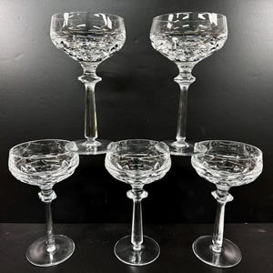 May include: Five clear crystal champagne glasses. The glasses have a round bowl with a textured pattern and a long stem. The glasses are arranged on a dark surface.