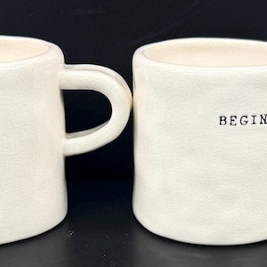 May include: Four off-white ceramic mugs with black text that reads "CALM.", "DREAM.", "BEGIN.", and "CREATE." Each mug has a rounded shape and a handle. The mugs are arranged in a row against a black background.