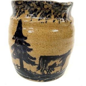May include: A tan ceramic pitcher with a dark blue rim and a rural scene painted on the side. The scene depicts a tree and a cow. The pitcher has a rustic, handmade appearance.