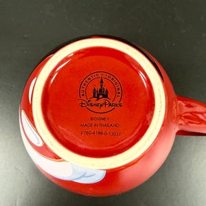 May include: A red ceramic mug with a white interior rim and a handle. The bottom of the mug features the Disney Parks logo, the words "AUTHENTIC ORIGINAL", and "MADE IN THAILAND".
