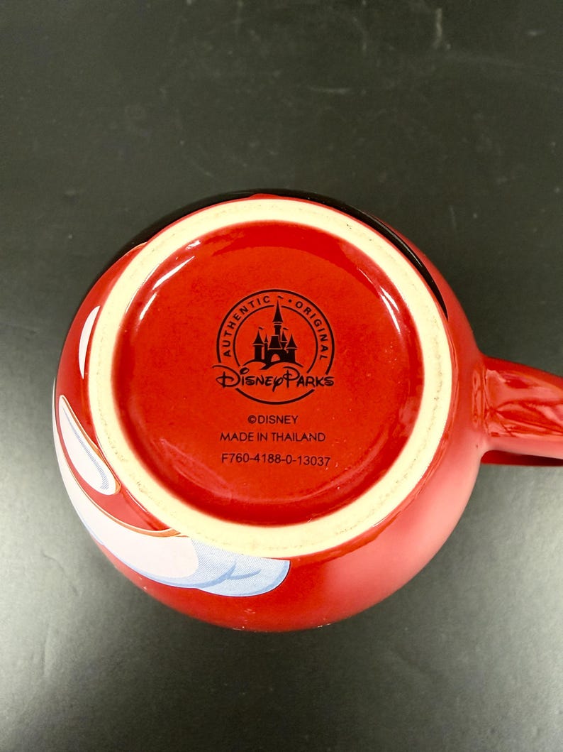 May include: A red ceramic mug with a white interior rim and a handle. The bottom of the mug features the Disney Parks logo, the words "AUTHENTIC ORIGINAL", and "MADE IN THAILAND".