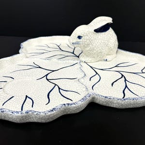 May include: A white ceramic serving dish with a bunny figurine. The dish is leaf-shaped with blue vein-like details and a blue trim. The bunny has blue accents on its ears and eyes.