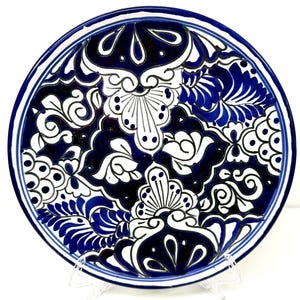 May include: A round ceramic plate featuring a bold, high-contrast pattern in deep cobalt blue and white. The design consists of intricate, symmetrical floral and scroll motifs, with solid blue sections and fine white line work throughout.