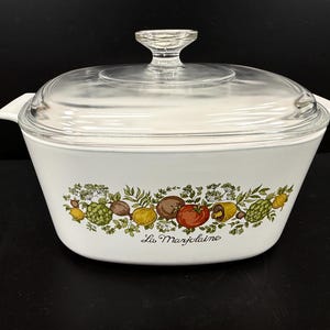 May include: White casserole dish with a clear glass lid and handle. The dish features a colorful vegetable design with the text "La Marjolaine" on the front. The dish has two white handles on either side.