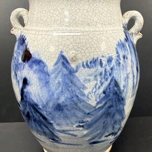 May include: A ceramic vase with a white crackle glaze finish and two side handles. The exterior features a hand-painted blue landscape design depicting stylized mountains and trees. The vessel has a wide mouth and a tapered base.