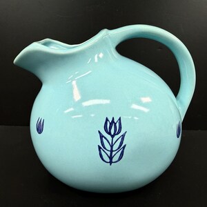 May include: A light blue ceramic pitcher with a rounded body and a curved handle. The pitcher features a spout for pouring and is decorated with dark blue tulip designs. The pitcher is approximately 8 inches tall.
