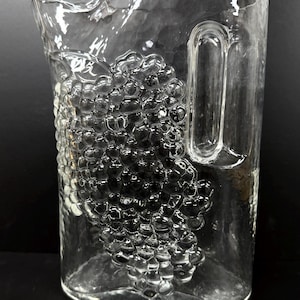 May include: A clear glass pitcher with a textured surface and a handle. A raised grape cluster design is featured on the front of the pitcher. The pitcher is designed for serving beverages.