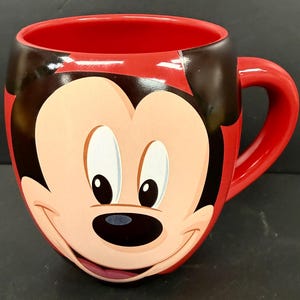 May include: Red ceramic mug featuring a smiling Mickey Mouse face. The mug has a red interior and handle, with black accents on the ears and around the face. A fun, collectible item for Disney fans.