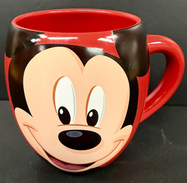 May include: Red ceramic mug featuring a smiling Mickey Mouse face. The mug has a red interior and handle, with black accents on the ears and around the face. A fun, collectible item for Disney fans.