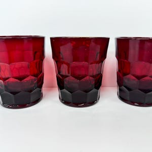 May include: Three ruby red drinking glasses with a geometric, faceted design. The glasses have a dark red base that fades to a lighter red towards the top. The glasses are made of a thick glass material.