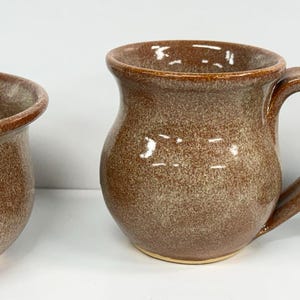 May include: Three handmade ceramic mugs in shades of brown and green. The mugs vary in shape and size, with one wide, shallow bowl-shaped mug, a rounded mug, and a taller mug with a dark green rim.