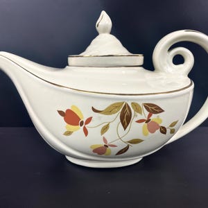 May include: A white teapot with a gold trim and a floral design. The teapot has a curved spout and a handle that curves back on itself. The floral design features brown, orange, and yellow leaves and flowers.