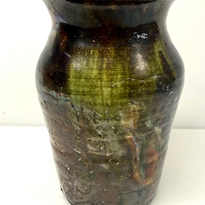 May include: A ceramic vase with a flared rim and a glossy, mottled glaze featuring deep shades of forest green, dark brown, and hints of metallic bronze. The surface has a textured, uneven finish with visible crackling and organic color variations.