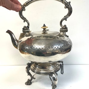May include: A silver teapot with an ornate handle and a stand with three legs. The teapot has a decorative design on the body and a small knob on the lid.