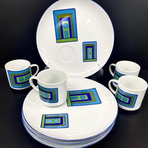 May include: Set of four white ceramic cups with blue and green geometric designs and a matching white ceramic serving plate with a blue rim. The cups have a handle and the plate has a raised edge.
