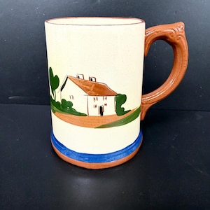 May include: A ceramic mug with a hand-painted landscape scene. The mug features a white house with a brown roof, green trees, and a brown handle. A blue stripe accents the base. The mug is approximately 6 inches tall.