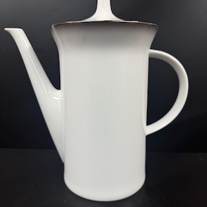 May include: White ceramic coffee pot with a curved spout and handle. The lid has a small, vertical handle. The rim of the lid has a dark brown accent. The pot is set against a black background.