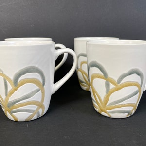 May include: Set of four white ceramic mugs with a gold and gray abstract design. The mugs have a rounded shape and a handle.