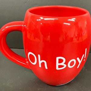 May include: A bright red ceramic mug with a large handle. The mug has the words "Oh Boy!" in white lettering on the side. The mug is glossy and appears to be empty.